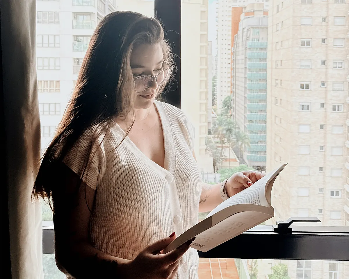 Ingryd Simões em pé, lendo um livro junto à janela, com prédios da cidade ao fundo